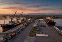 El agro normaliza los fletes en casi todo el país, pero un bloqueo en Quequén frena la salida de granos Una vista aérea al atardecer de un puerto granario que está parcialmente bloqueado. Una fila de camiones está detenida y personas sostienen una pancarta vacía en el muelle frente a barcos de carga amarrados.