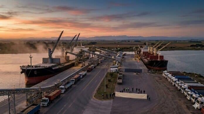 Una vista aérea al atardecer de un puerto granario que está parcialmente bloqueado. Una fila de camiones está detenida y personas sostienen una pancarta vacía en el muelle frente a barcos de carga amarrados.