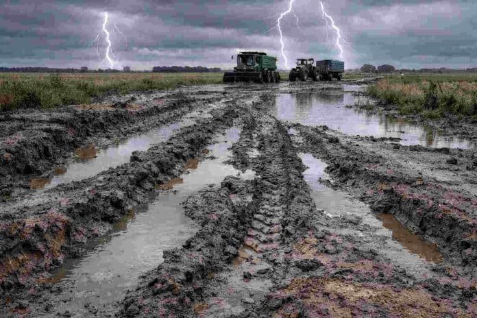 Camino rural intransitable con barro y agua acumulada tras lluvias intensas, con maquinaria agrícola detenida en el fondo