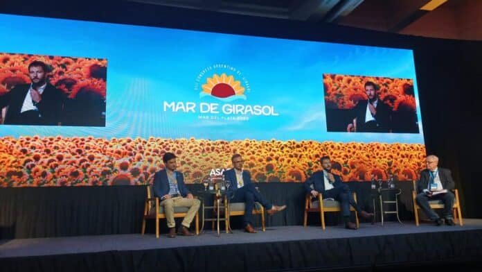 Panel sobre comercio global del girasol en el Congreso Argentino de Girasol en Mar del Plata, con expositores en escenario y pantalla con campo de girasoles de fondo.