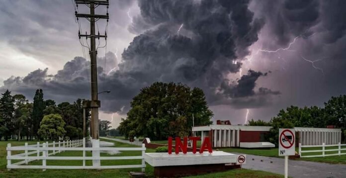 inta-tormenta-cielo-amenazante-1200px-ruralnet Entrada del INTA con cartel institucional bajo cielo oscuro de tormenta, nubes cumulonimbus y relámpago en el fondo, ambiente lluvioso y amenazante en un predio rural argentino.