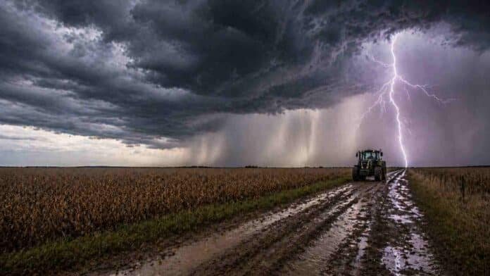 Tractor avanzando por un camino rural embarrado bajo una tormenta con lluvia intensa y un relámpago fuerte en el horizonte, reflejando condiciones climáticas adversas en el campo argentino.