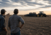 La confianza del campo cayó 16% en dos meses: costos en alza, inversiones frenadas y cautela para sembrar trigo Dos productores rurales conversan de espaldas frente a un campo en barbecho con tractor y sembradora detenidos, reflejando incertidumbre económica y caída de la confianza en el agro argentino.
