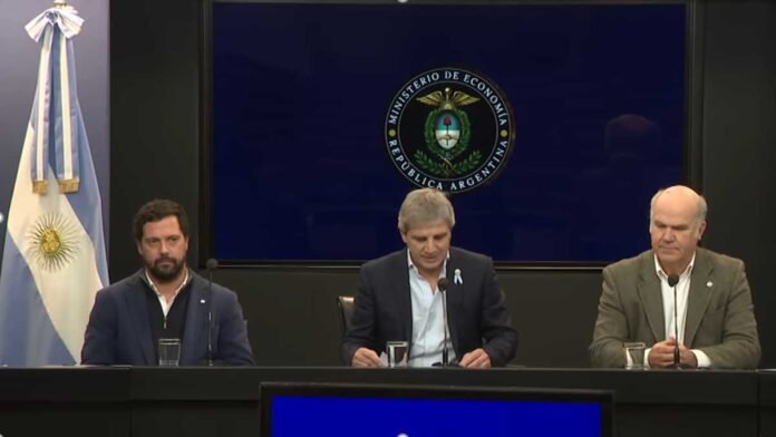 Conferencia económica en el Ministerio de Economía Funcionarios argentinos brindan una conferencia de prensa en el Ministerio de Economía. Tres hombres sentados frente a micrófonos aparecen bajo el escudo oficial de la República Argentina, con una bandera argentina al costado, en una imagen institucional de formato panorámico.