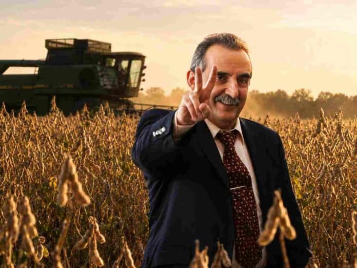 Dirigente rural sonríe y hace la V en un lote de soja durante la cosecha, con una cosechadora trabajando al fondo al atardecer.