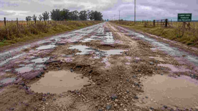 Camino rural en muy mal estado con barro, pozos profundos y charcos de agua, rodeado de campos y alambrados bajo un cielo nublado.