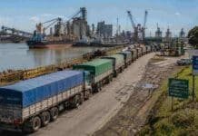 Santa Fe defiende el fideicomiso portuario: “A nadie le gusta pagar, pero las rutas no aguantan más Fila de camiones cerealeros avanzando hacia una terminal portuaria argentina junto al río, con barcos, grúas y plantas industriales visibles en el puerto.