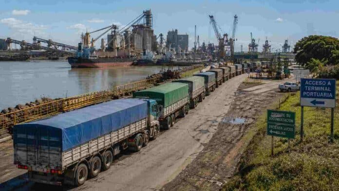 Fila de camiones cerealeros avanzando hacia una terminal portuaria argentina junto al río, con barcos, grúas y plantas industriales visibles en el puerto.
