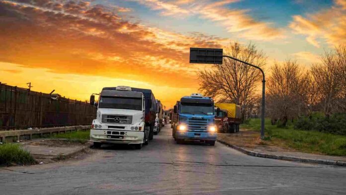 Fila de camiones cargados con granos esperando ingresar a terminales portuarias del Gran Rosario bajo un cielo de atardecer.