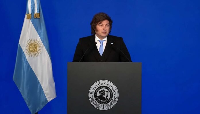discurso-presidente-bolsa-cereales-argentina-ruralnet Dirigente político argentino durante un discurso en la Bolsa de Cereales, con bandera nacional y fondo azul en un acto institucional.