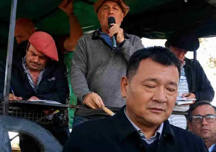 Productores y representantes rurales durante un remate ganadero, con un hombre hablando por micrófono y asistentes tomando nota en la tribuna.