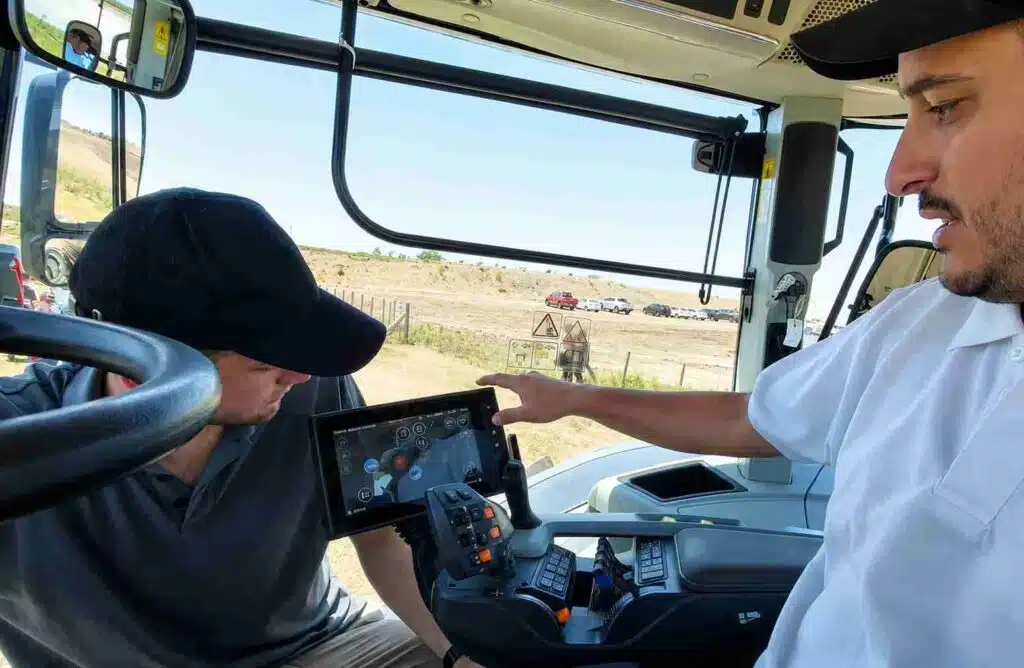 Interior de la cabina de una maquinaria agrícola durante una experiencia de manejo, con un instructor señalando la pantalla de control al operador.