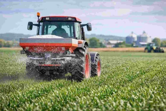 fertilizacion_cultivos “Tractor rojo aplicando fertilizante granular sobre un cultivo verde bajo cielo parcialmente nublado, en formato 16:9.”