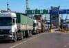 Los números que muestran el verdadero peso del campo en la economía Fila de camiones cerealeros esperando ingreso a una terminal portuaria argentina, con grúas y estructuras industriales al fondo en una escena agroexportadora.