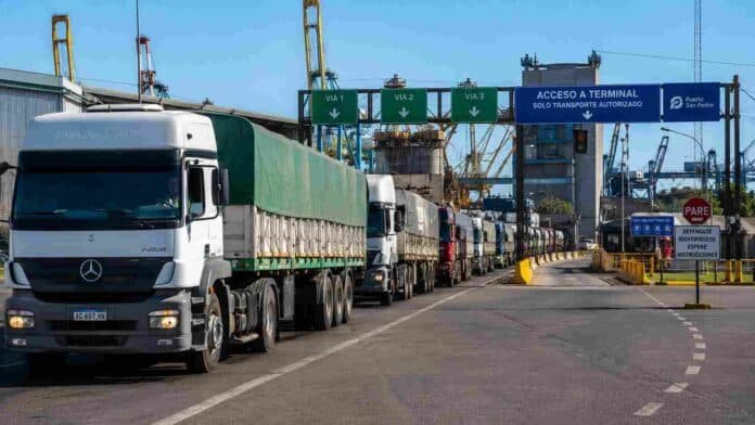 fila-camiones-acceso-portuario-granos-ruralnet Fila de camiones cerealeros esperando ingreso a una terminal portuaria argentina, con grúas y estructuras industriales al fondo en una escena agroexportadora.