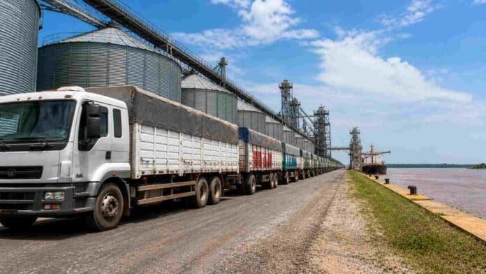 Fila de camiones cargados con granos esperando descarga en puerto agroexportador argentino, con silos y barco granelero a la vera del río.