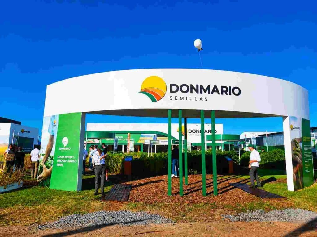 Stand institucional de Don Mario Semillas en una exposición agropecuaria, con visitantes recorriendo el espacio y parcelas demostrativas de cultivos.