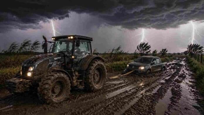 Tractor Valtra embarrado tirando con una linga de una camioneta Amarok encajada en un camino rural con barro, bajo tormenta intensa con relámpagos y lluvias en el Litoral argentino.