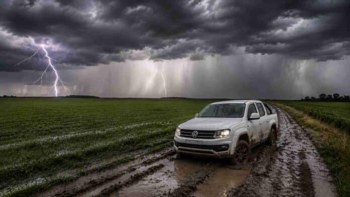 camioneta-encajada-camino-rural-tormenta-electrica Camioneta blanca encajada en un camino rural embarrado durante una tormenta severa, con lluvias intensas, relámpagos y un campo agrícola bajo un cielo oscuro y amenazante.