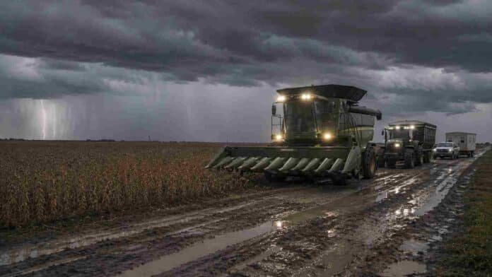Cosechadora sojera con luces encendidas avanza por un camino rural embarrado con huellones y charcos junto a un lote de soja en estado R7 bajo una tormenta con lluvia y relámpagos en la región núcleo argentina.