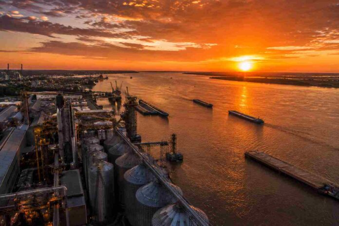 Vista aérea del puerto de Rosario al atardecer, con silos y elevadores de granos junto al río Paraná y barcazas navegando bajo un cielo dorado.