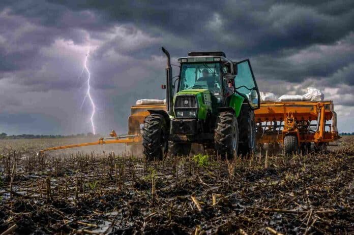 Tractor con sembradora trabajando sobre suelo húmedo con rastrojo bajo cielo de tormenta y un relámpago en el horizonte.