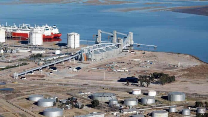 Vista aérea de una terminal portuaria e industrial junto a un río, con silos, tanques de almacenamiento y buques de carga destinados a exportación.