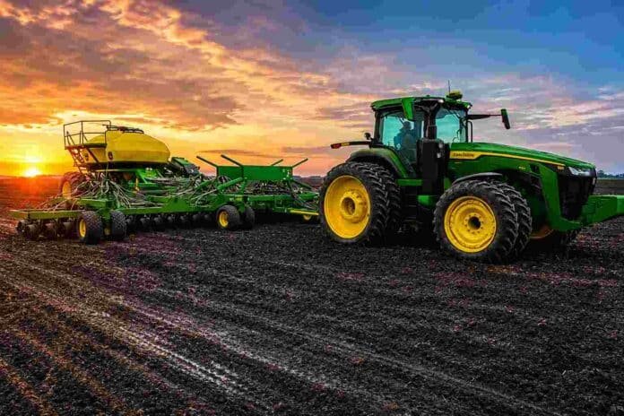 Tractor con sembradora sobre campo en barbecho y suelo húmedo al atardecer, con cielo de colores intensos en una escena agrícola