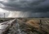 Avanzan lluvias, heladas y ráfagas: el pronóstico completo para el campo Camino rural en campo abierto con molino, pastizales y cielo de tormenta, mientras avanza una cortina de lluvia intensa sobre el horizonte.
