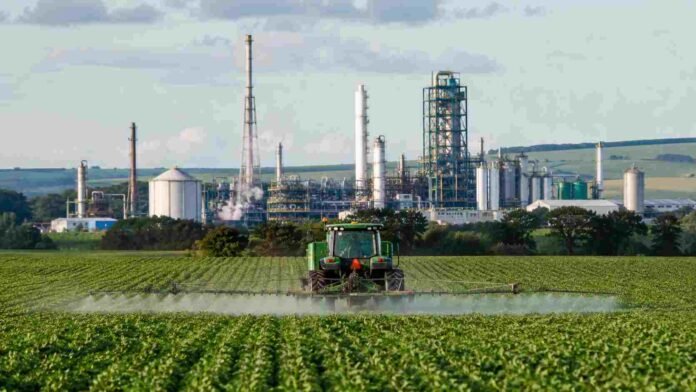 Tractor aplicando fertilizantes en un campo agrícola de Brasil, con una planta industrial de urea y fertilizantes al fondo.