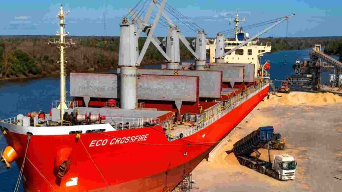 Buque de carga en un puerto fluvial durante la operatoria de granos, con camión descargando cereal y maquinaria portuaria.