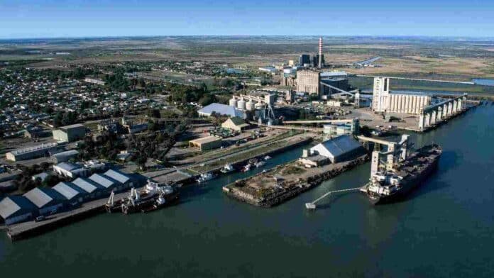 Vista aérea de un puerto industrial con silos, terminales de carga, depósitos y buques amarrados.
