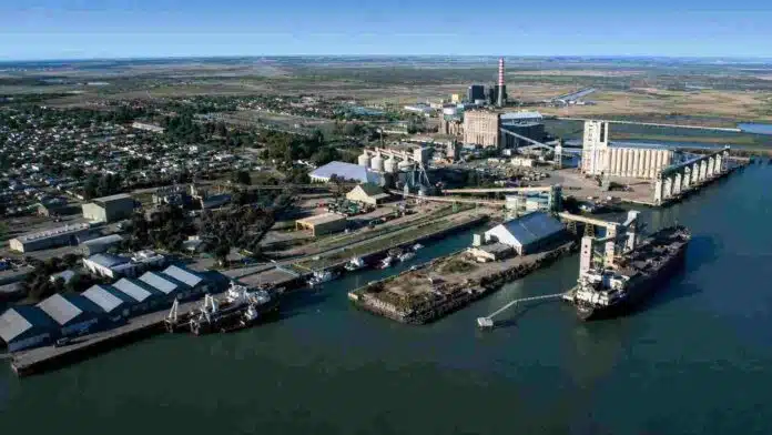 Vista aérea de un puerto industrial con silos, terminales de carga, depósitos y buques amarrados.