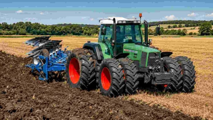 Tractor agrícola verde con arado trabajando sobre un lote cosechado, removiendo el suelo en un campo rural bajo cielo despejado.