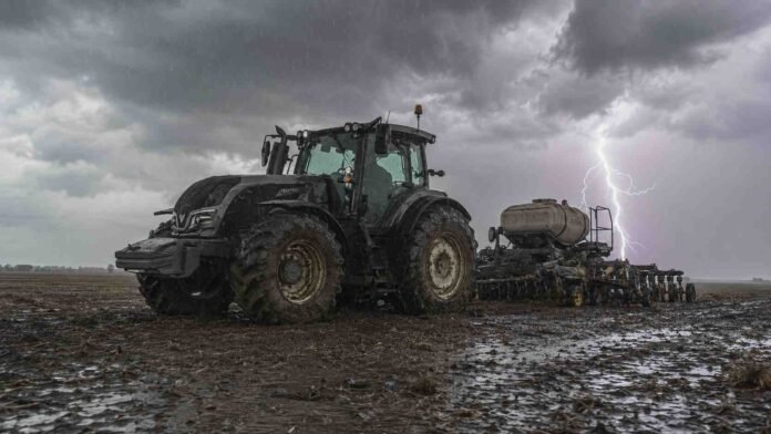 tractor-valtra-sembradora-pierobon-lluvia-relampago-ruralne Tractor Valtra con sembradora Pierobon de granos finos trabajando en un campo en barbecho, con suelo mojado, lluvia y un relámpago bajo cielo nublado.