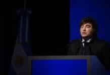 Milei defendió el rumbo económico y destacó el crecimiento Orador durante un discurso en un acto oficial, frente a un atril y con la bandera argentina de fondo.