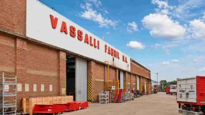 fabrica-vassalli-firmat-santa-fe Fachada de la fábrica Vassalli en Firmat, Santa Fe, con maquinaria y vehículos en el predio industrial bajo un cielo azul con nubes.