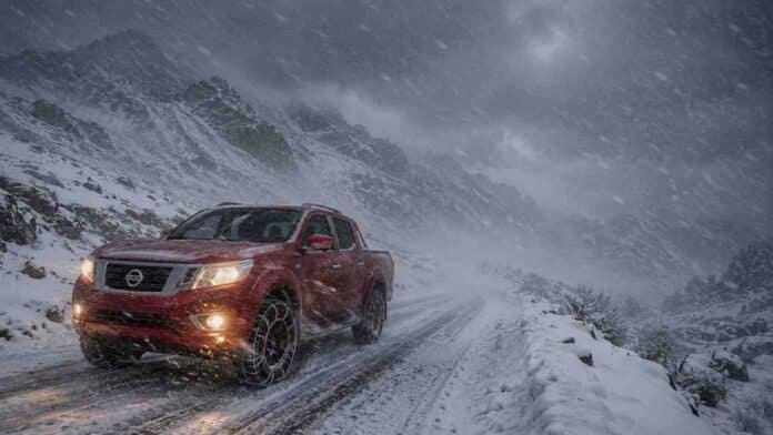 Pickup 4x4 roja con cadenas en los neumáticos circulando por una ruta de montaña nevada en la cordillera patagónica, con viento blanco y baja visibilidad.
