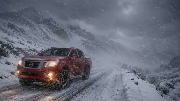 Pickup 4x4 roja con cadenas en los neumáticos circulando por una ruta de montaña nevada en la cordillera patagónica, con viento blanco y baja visibilidad.