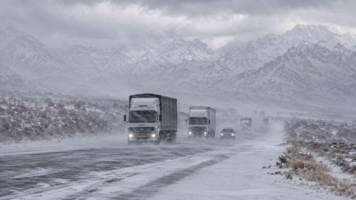 ruta-nevada-camiones-vehiculos-cordillera-mendoza-2026 Camiones y vehículos circulan por una ruta nevada de Mendoza bajo viento blanco, con la cordillera de los Andes cubierta de nieve al fondo.