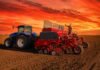 Siembra de trigo: el avance llegó al 99,8% y persisten excesos de humedad Tractor azul y sembradora roja trabajando en un campo agrícola durante un atardecer de tonos rojizos