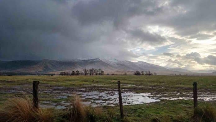 Campo húmedo con charcos bajo un cielo de tormenta y sierras con nieve, por el pronóstico agropecuario para Buenos Aires.