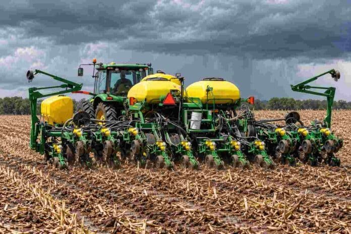Tractor con sembradora de maíz trabajando sobre rastrojo y suelo húmedo bajo un cielo cubierto por nubes de lluvia.