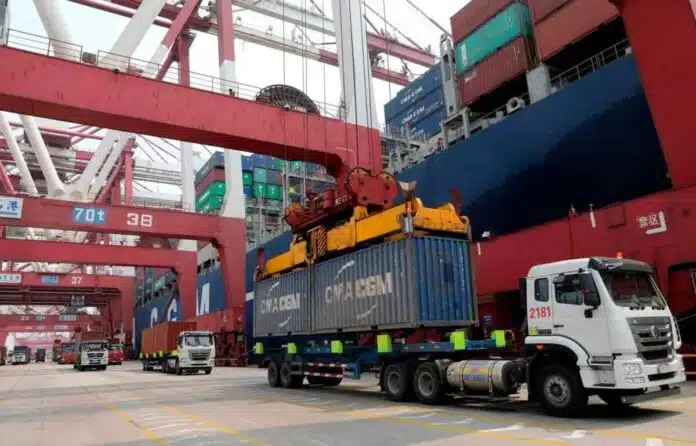 exportaciones-comercio-exterior-puerto-contenedores Camiones transportan contenedores de carga en una terminal portuaria, mientras grúas de gran porte operan junto a un buque de carga en el marco del comercio internacional.