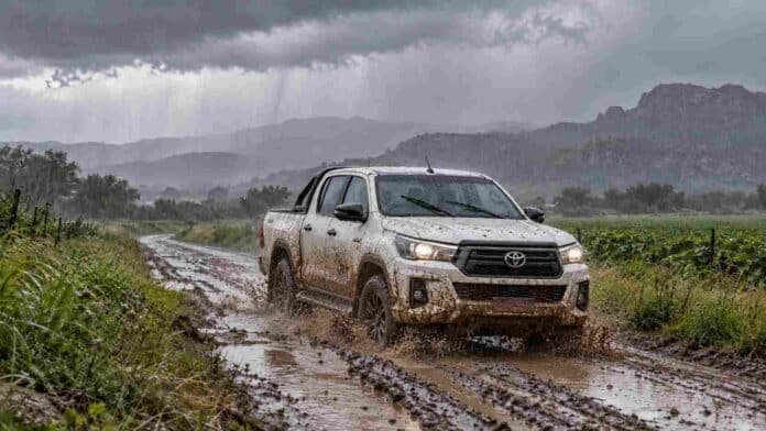 Camino rural embarrado bajo la lluvia con una camioneta Hilux blanca salpicada de barro avanzando entre charcos, en un paisaje agropecuario con tormenta.