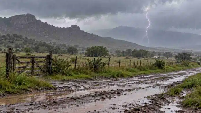 lluvias-persistentes-cordoba-la-rioja-san-luis-ruralnet Lluvias intensas sobre un paisaje rural serrano, con camino de tierra inundado, charcos y un relámpago bajo nubes de tormenta.