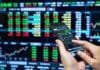 El Mercado Argentino de Valores marcó un récord de operaciones en 2026 Persona operando desde un teléfono móvil frente a pantallas con cotizaciones y gráficos del mercado financiero.