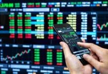 El Mercado Argentino de Valores marcó un récord de operaciones en 2026 Persona operando desde un teléfono móvil frente a pantallas con cotizaciones y gráficos del mercado financiero.