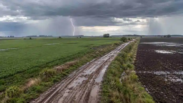 pronostico-lluvias-tiempo-inestable-15-17-octubre-ruralnet Vista aérea de campos agrícolas y camino rural embarrado, con lluvias y un relámpago bajo un cielo de tormenta en la región pampeana argentina.